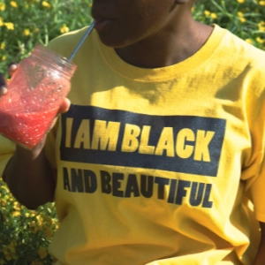 femme noire dans les champs buvant un smoothie rouge et portant un t-shirt jaune avec floqué dessus "I am black and beautiful"