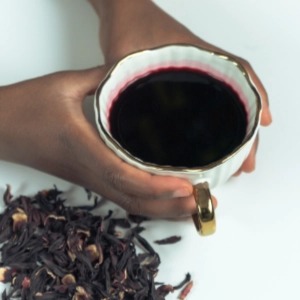 mains d'une femme noire tenant une tasse remplie de bissap et avec des fleurs d'hibiscus séchées sur la table